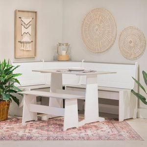 A white dining table with benches sits on a patterned rug, surrounded by neutral walls, decorative wall hangings, a plant, and a lit lantern in a cozy setting.