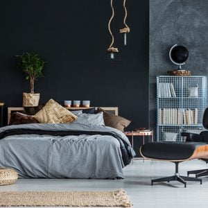 A bed with layered bedding sits in a stylish room. Soft lighting and decor create a cozy atmosphere, complemented by furniture and a dark wall.