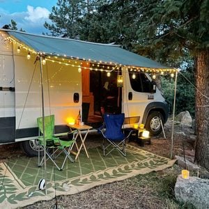 A camper van stands in a natural setting, illuminated by string lights, with two chairs and a small table placed on a decorative rug.