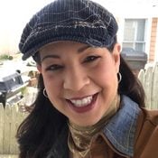A woman wearing a dark cap smiles brightly. She stands in front of a wooden fence, with an outdoor grill and house visible in the background.