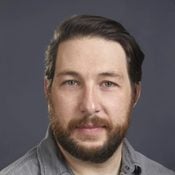 A man with dark hair and a beard faces the camera, wearing a gray shirt against a neutral background. His expression appears calm and focused.