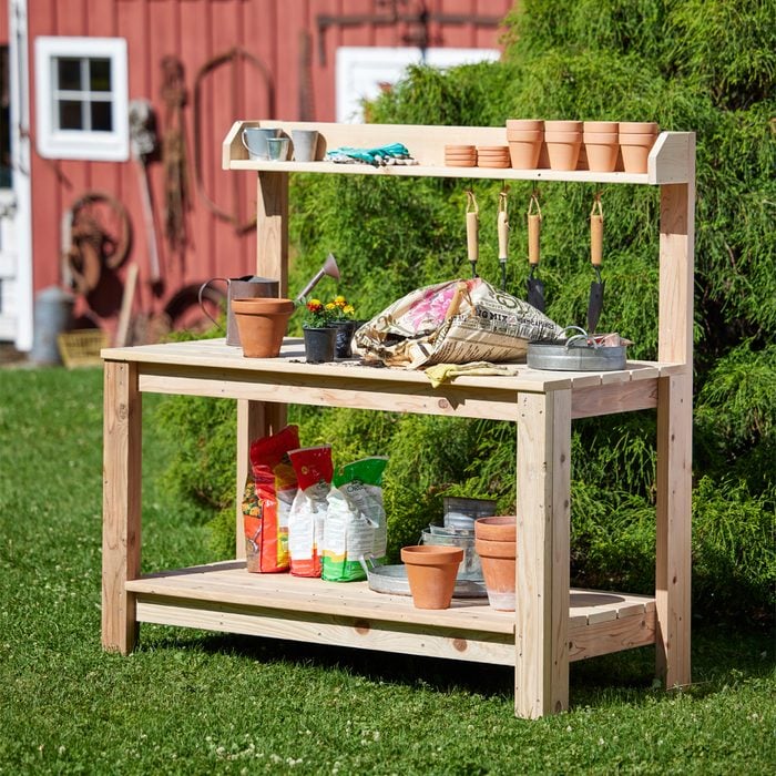 A wooden potting table displays gardening tools, pots, soil bags, and flowers outdoors, surrounded by green grass and a red barn in the background.