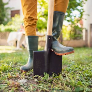 A person in green boots stands on a shovel in a garden, preparing to dig into the grass surrounded by greenery and a house in the background.
