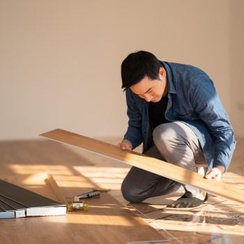A man examines a wooden plank while kneeling on the floor. Tools and additional planks are nearby in a bright, empty room.