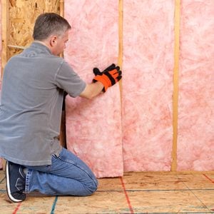 A man kneels, installing pink insulation between wooden studs in a construction space, wearing gloves and jeans on an unfinished floor.