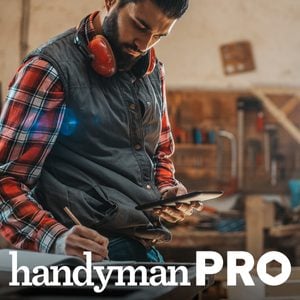 A man wearing safety headphones reviews plans on a tablet while seated at a workbench in a workshop filled with tools and woodwork materials.