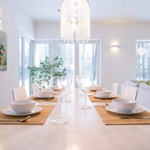 A dining table is set elegantly with white dishes and glassware, illuminated by a chandelier, surrounded by large windows revealing a snowy landscape.