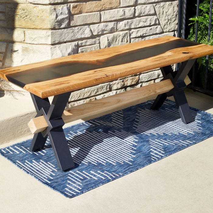 A wooden bench with a sleek design sits on a patterned blue rug, beside a stone wall and greenery, creating a cozy outdoor setting.